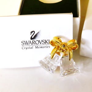 May include: A Swarovski Crystal Memories ornament featuring two clear, faceted crystal bells suspended from a polished gold-tone metal bow. The item is displayed in front of a white card with the black Swarovski swan logo and the text Crystal Memories.