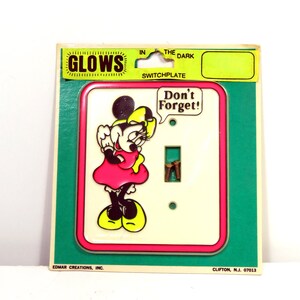 May include: A light switch plate featuring a cartoon image of Minnie Mouse in a pink dress and yellow shoes. The plate is white with a pink border and the text "Don't Forget!" in a speech bubble above Minnie Mouse. The plate glows in the dark.