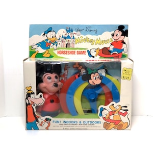 May include: A vintage Walt Disney Mickey Mouse horseshoe game in a box. The box features a colourful illustration of Mickey Mouse, Donald Duck, and Goofy. The game includes a red, yellow, blue, and green horseshoe set with a Mickey Mouse figure in the centre.
