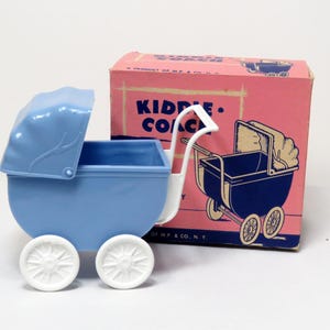 May include: A vintage blue toy baby carriage with white wheels and a white handle. The carriage is next to a pink box with the words "Kiddie Coach" printed on it. The box has a drawing of a baby carriage.