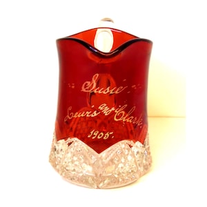 May include: A ruby red glass pitcher featuring a clear, faceted base with a diamond-cut pattern. The body of the pitcher is engraved with the script text "Susie", "Lewis and Clark", and the year "1905" in a light, etched finish.