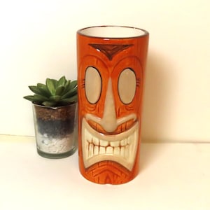 May include: Orange tiki-style ceramic tumbler with a smiling face design. The tumbler features a detailed tiki mask with large eye shapes, a prominent nose, and a wide, toothy grin. A small succulent plant in a glass container is next to the tumbler.