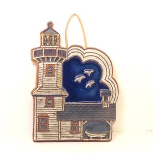 May include: A ceramic wall plaque featuring a lighthouse and an adjacent building. The lighthouse is white with dark blue accents, while the sky is a deep glossy blue with three white birds. A tan cord is attached at the top for hanging purposes.