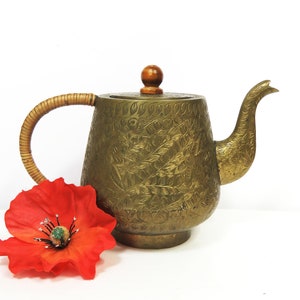 May include: A gold-colored brass teapot with an ornate floral design and a wooden knob on the lid. The teapot has a woven handle and is sitting on a white surface.