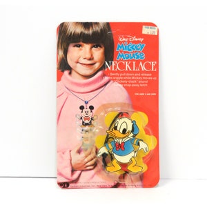 May include: A vintage Walt Disney Mickey Mouse necklace in original packaging. The red card features a child wearing the necklace and text describing the toy's movement and sound. A Donald Duck figure is visible inside the clear plastic blister pack.