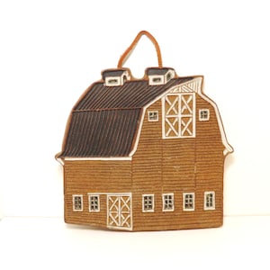May include: A brown ceramic barn-shaped ornament with a dark brown roof and white accents. The barn features windows, doors, and a leather strap for hanging. A decorative piece for country-themed decor.