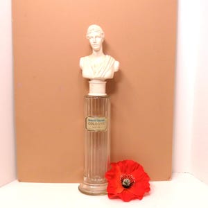 May include: A vintage cologne bottle with a white bust of a woman on top. The clear glass bottle has vertical ridges and a label that reads "Cologne". A bright red poppy flower rests near the bottle.