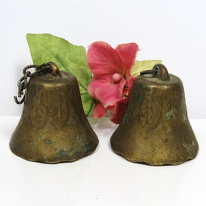 May include: Two antique brass bells with a weathered patina and a subtle embossed leaf pattern around the top. Each bell features a small metal loop at the crown for hanging, with one bell retaining a short segment of a metal chain link.