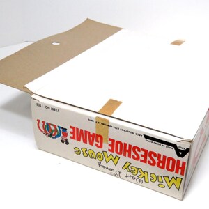 May include: A cardboard box for a Mickey Mouse Horseshoe Game. The box is red, white, and blue with a picture of Mickey Mouse and his friends. The text on the box reads "Mickey Mouse Horseshoe Game" and "Item No. 1106".
