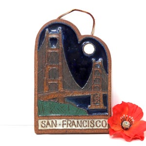 May include: A brown ceramic heart-shaped ornament with a blue and green glaze depicting the Golden Gate Bridge in San Francisco. The ornament has a hole at the top for hanging and reads "SAN FRANCISCO" at the bottom.