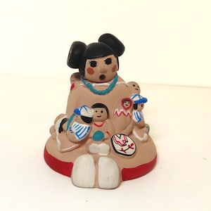 May include: A hand-painted ceramic figurine of a woman with black hair in two buns, wearing a turquoise necklace, and surrounded by several small children. The figurine has a tan body, red base, and white accents.