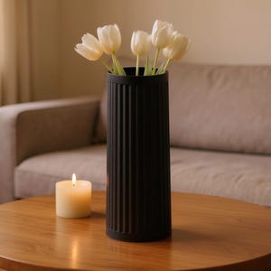 Op de afbeelding: Een zwarte, geribbelde vaas met witte tulpen staat op een houten tafel. Een brandende kaars staat naast de vaas. De tafel staat voor een beige bank, wat een gezellige woondecoratie creëert.