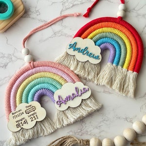 May include: Two handmade rainbow wall hangings with macrame fringe. One rainbow has the name "Andrew" on a cloud-shaped wooden cutout. The other has "Amalia" and birth details: 8 lbs 14 oz, 21 in, born 7/11/24 at 12:31 PM.