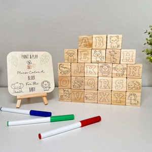 May include: Wooden alphabet blocks with animal illustrations and letters A-Z. A small wooden sign reads "Paint & Play, Please Color A Block For The Baby." Markers in red, green, blue, and yellow are included.