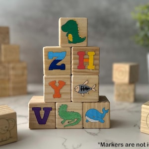 May include: Wooden alphabet blocks with colorful illustrations of a dinosaur, fish, iguana, and whale. The blocks also feature the letters Z, H, Y, V, and C. The text "*Markers are not included" is at the bottom.