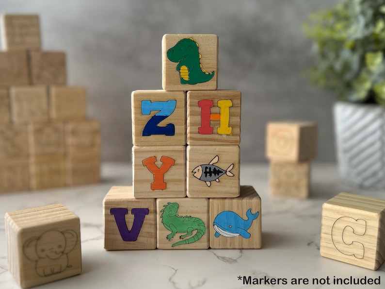 May include: Wooden alphabet blocks with colorful illustrations of a dinosaur, fish, iguana, and whale. The blocks also feature the letters Z, H, Y, V, and C. The text "*Markers are not included" is at the bottom.