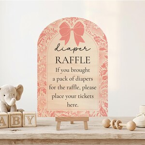 May include: A wooden sign with the text "diaper RAFFLE" and a pink bow design. The sign is on a small easel, with a stuffed elephant, wooden blocks spelling "BABY", and baby shoes nearby. A baby shower theme.