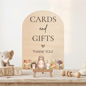 May include: Wooden sign with the words "CARDS and GIFTS" and "THANK YOU!" in black script. The sign features a watercolor illustration of woodland animals. A plush elephant, wooden blocks spelling "BABY", and baby shoes are also visible.
