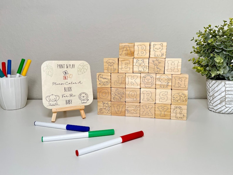 May include: Wooden alphabet blocks with animal illustrations and letters A-Z. A small wooden sign reads "Paint & Play, Please Color A Block For The Baby." Markers in red, green, blue, and yellow are included.