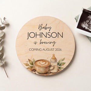 May include: A round wooden sign with the text "Baby JOHNSON is brewing COMING AUGUST 2026". The sign features a watercolor illustration of a coffee cup with a heart design, a baby bottle, and coffee beans. An ultrasound image is also visible.