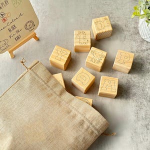 May include: Wooden alphabet blocks with animal illustrations and letters, alongside a small easel with text that reads "Paint & Play, Please Color A Block For The Baby". A beige drawstring bag is also present.