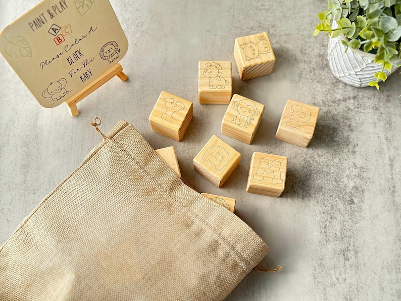 May include: Wooden alphabet blocks with animal illustrations and letters, alongside a small easel with text that reads "Paint & Play, Please Color A Block For The Baby". A beige drawstring bag is also present.