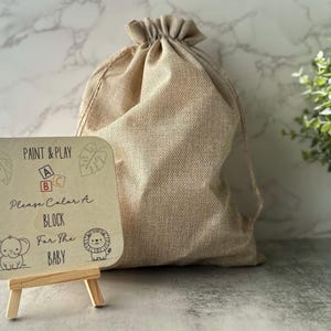 May include: A beige burlap drawstring bag next to a small wooden easel holding a sign. The sign reads "Paint & Play" with block letters and illustrations of an elephant and lion. The text on the sign says "Please Color A Block For The Baby."