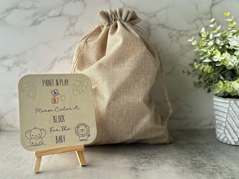 May include: A beige burlap drawstring bag next to a small wooden easel holding a sign. The sign reads "Paint & Play" with block letters and illustrations of an elephant and lion. The text on the sign says "Please Color A Block For The Baby."