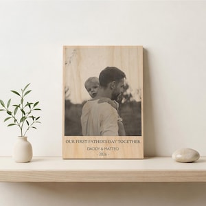 May include: A rectangular wooden plaque featuring a black and white photograph of a parent holding a child. The text reads: OUR FIRST FATHER'S DAY TOGETHER, DADDY & MATTEO, 2026. The natural wood grain is visible throughout the surface of the plaque.