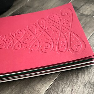 May include: A stack of red Christmas cards with a white embossed tree design. The tree is decorated with swirls and stars.