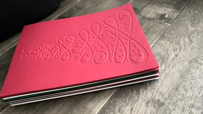 May include: A stack of red Christmas cards with a white embossed tree design. The tree is decorated with swirls and stars.