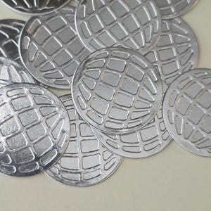 May include: A collection of silver-colored, circular metal charms. Each charm features a raised grid pattern, resembling a globe. The charms are scattered across a light-colored surface, creating a textured and reflective display.