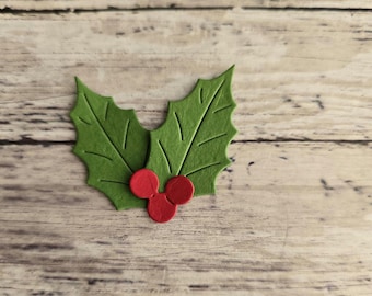 May include: A green holly leaf with three red berries. The holly is made of felt and is set against a wooden background. The leaves have detailed veining.