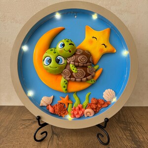 May include: A circular artwork with a light-up frame. It depicts two turtles on an orange moon, with a yellow star and sea life details. The background is blue. The frame has small lights around it, adding to the decorative effect.