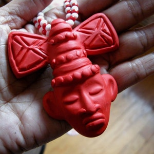 May include: A red pendant necklace featuring a stylized face with closed eyes and a decorative headpiece. The pendant is suspended from a beaded necklace with alternating red and white beads. The pendant is held in a hand.