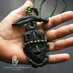 May include: Black pendant necklace featuring a stylized face with a headdress and green accents. The necklace has a black cord with green beads. The pendant is handmade and has the text "OLGA AYALA HANDICRAFTS" and "(Hecho A Mano)".