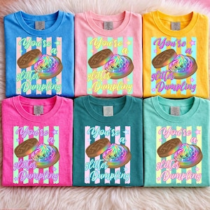 Puede incluir: Seis camisetas coloridas con el texto "You're a glitter Dumpling" y una ilustración de una empanadilla. Las camisetas son de color azul, rosa, amarillo, verde azulado y verde.