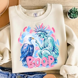 May include: Cream-coloured sweatshirt with a graphic of the Statue of Liberty touching the beak of a blue and white eagle. The word "BOOP" is written in pink. The background has pink and blue brushstrokes.