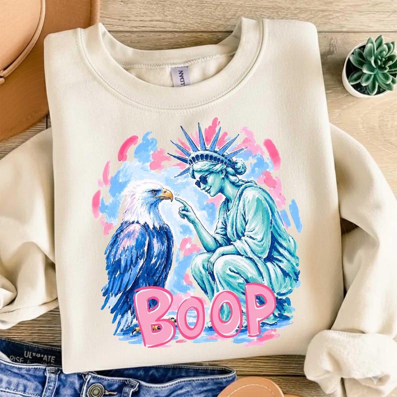 May include: Cream-coloured sweatshirt with a graphic of the Statue of Liberty touching the beak of a blue and white eagle. The word "BOOP" is written in pink. The background has pink and blue brushstrokes.