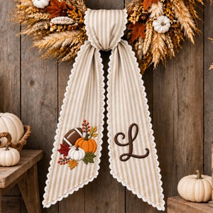 May include: A beige and white striped fabric ribbon with white lace trim, featuring an embroidered football, orange and white pumpkins, and autumn leaves on one side, and a large brown embroidered letter L on the other, hanging from a wreath.