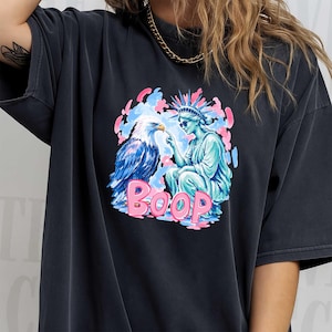 May include: Dark grey t-shirt with a graphic of an eagle and the Statue of Liberty, and the word "BOOP" in pink. The eagle is blue and white, and the Statue of Liberty is turquoise. The background has pink and blue accents.