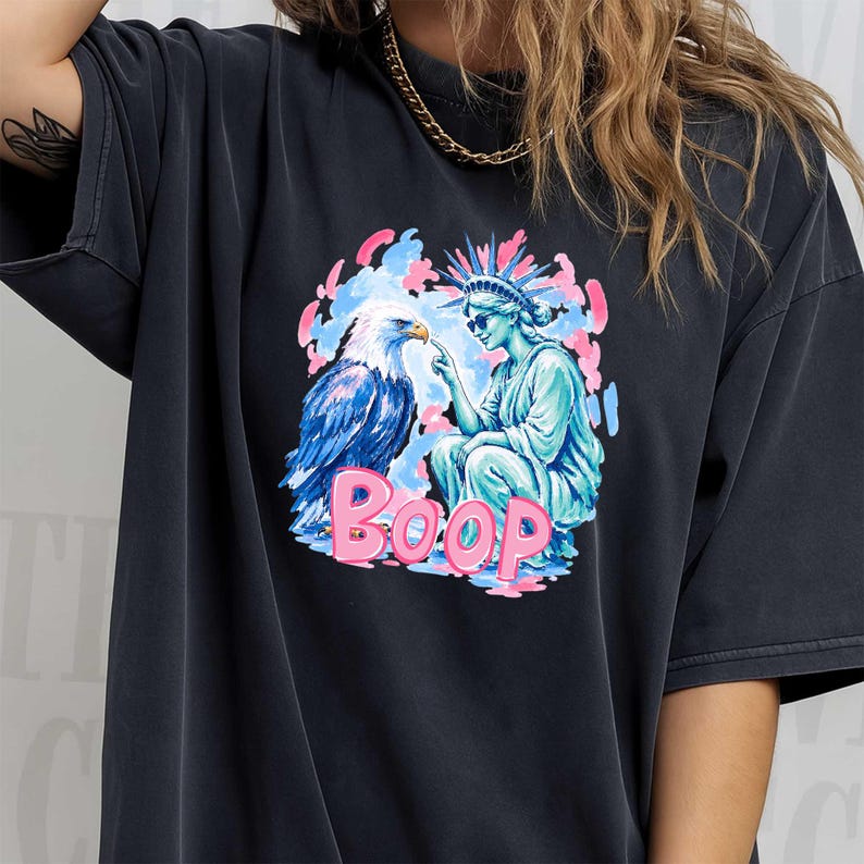 May include: Dark grey t-shirt with a graphic of an eagle and the Statue of Liberty, and the word "BOOP" in pink. The eagle is blue and white, and the Statue of Liberty is turquoise. The background has pink and blue accents.