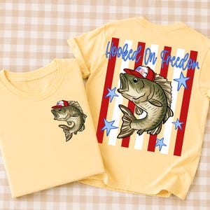 May include: Two yellow t-shirts with a patriotic theme. One shirt features a small graphic of a fish wearing a red, white, and blue hat. The other shirt has a larger graphic of the same fish, red and white stripes, blue stars, and the text "Hooked On Freedom."