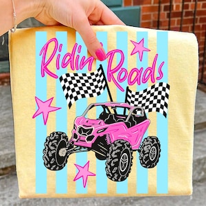May include: A yellow towel with blue vertical stripes and a pink off-road vehicle graphic. The text "Ridin' Roads" is in pink script. Two checkered flags and pink stars complete the design.