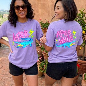Könnte beinhalten: Zwei Personen tragen lavendelfarbene T-Shirts mit Alligatoren-Grafiken. Auf einem Shirt steht "Later" und auf dem anderen "After While". Das Alligator ist blau und gelb mit einem grünen Blitz. Sie tragen schwarze Shorts.