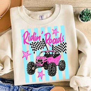 Peut inclure: Sweat-shirt cr&egrave;me avec un motif repr&eacute;sentant un v&eacute;hicule tout-terrain rose, des drapeaux &agrave; damiers et le texte "Ridin' Roads" en rose. Le motif comprend des rayures bleu clair et des &eacute;toiles roses. C'est un v&ecirc;tement d&eacute;contract&eacute;.