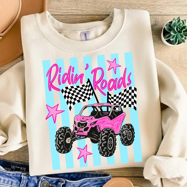 Peut inclure: Sweat-shirt cr&egrave;me avec un motif repr&eacute;sentant un v&eacute;hicule tout-terrain rose, des drapeaux &agrave; damiers et le texte "Ridin' Roads" en rose. Le motif comprend des rayures bleu clair et des &eacute;toiles roses. C'est un v&ecirc;tement d&eacute;contract&eacute;.