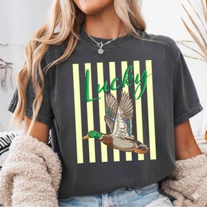 May include: Dark gray t-shirt featuring a flying duck illustration with green, brown, and blue details. The word "Lucky" is written in green script above the duck, set against a background of yellow vertical stripes.