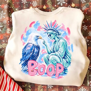 May include: Cream-coloured sweatshirt with a graphic of the Statue of Liberty, an eagle, and the word "BOOP" in pink. The Statue of Liberty is blue, wearing sunglasses and pointing at the eagle.