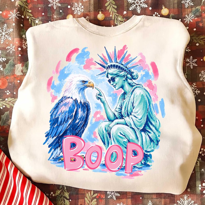 May include: Cream-coloured sweatshirt with a graphic of the Statue of Liberty, an eagle, and the word "BOOP" in pink. The Statue of Liberty is blue, wearing sunglasses and pointing at the eagle.